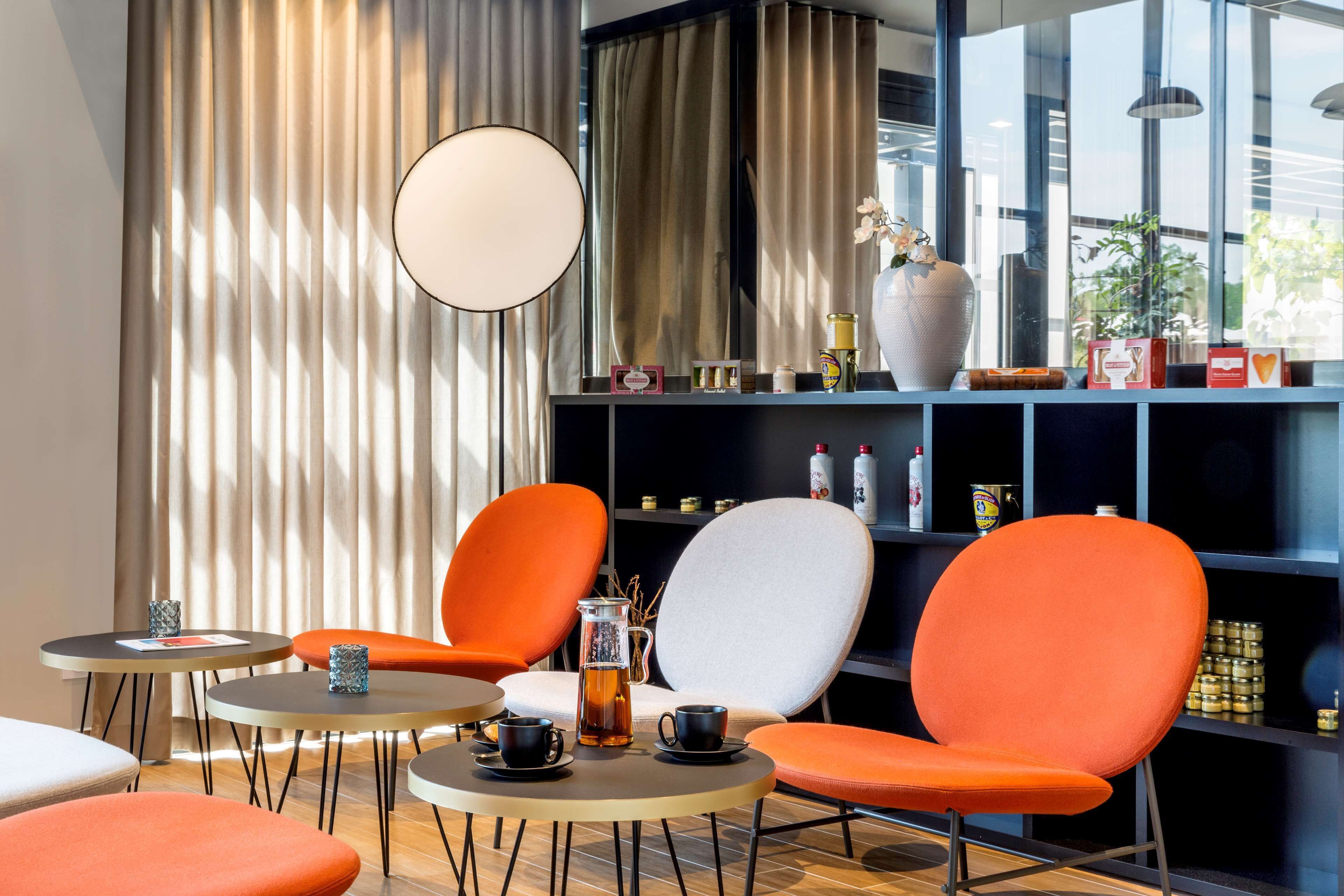 Ein modernes Interieur mit runden Tischen, orangefarbenen Stühlen, einer großen Vase und einem Regal mit diversen Gegenständen.