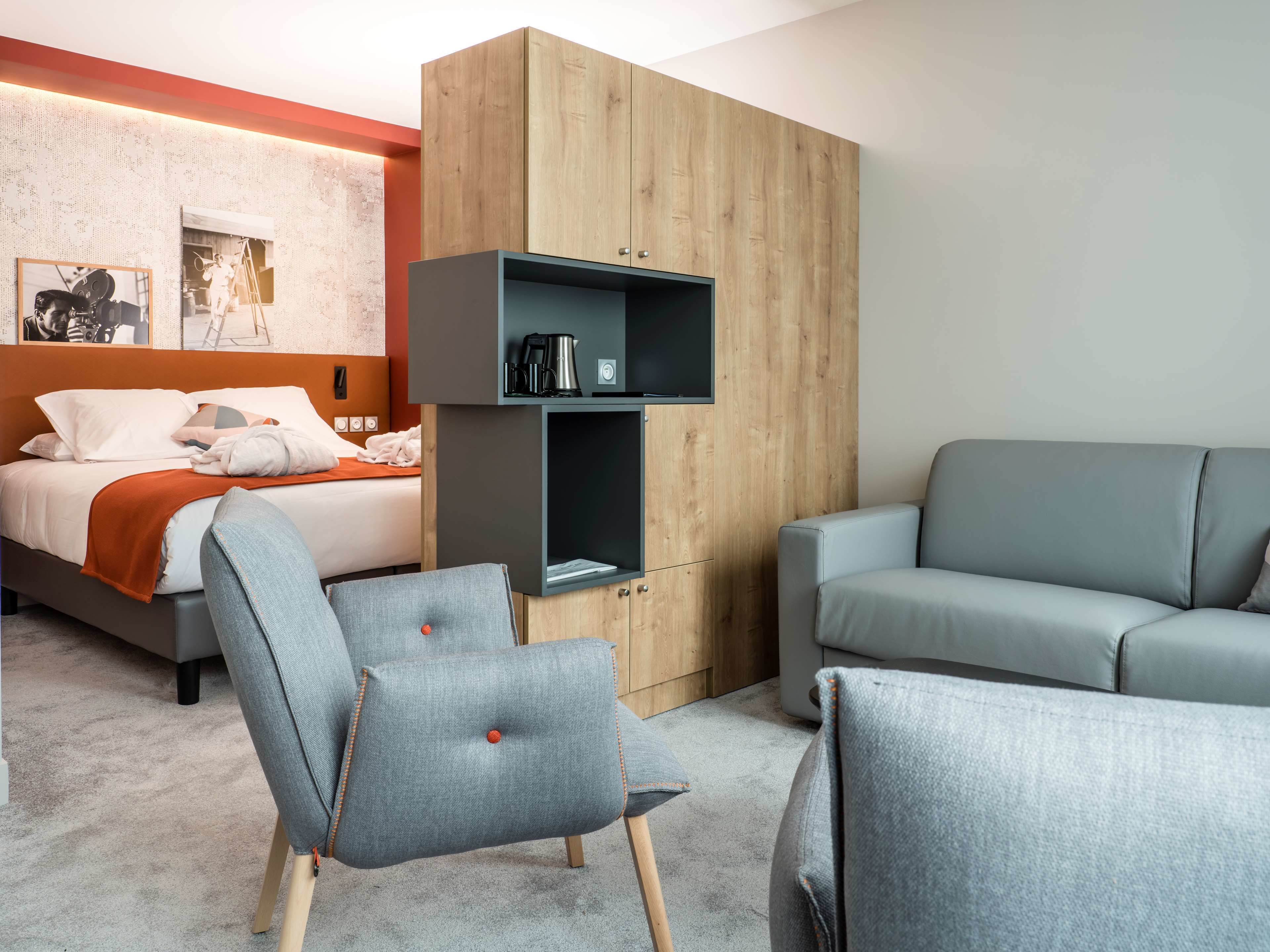 A modern hotel room with a bed, grey upholstered chairs, a wooden wardrobe, and a flat-screen TV.
