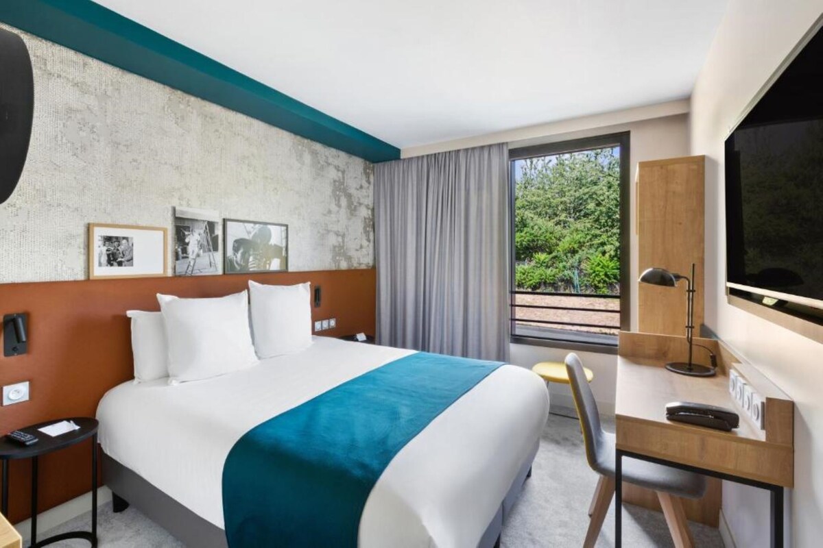 A hotel room with a large bed, a desk with a chair, a TV, and a window with a view of greenery.