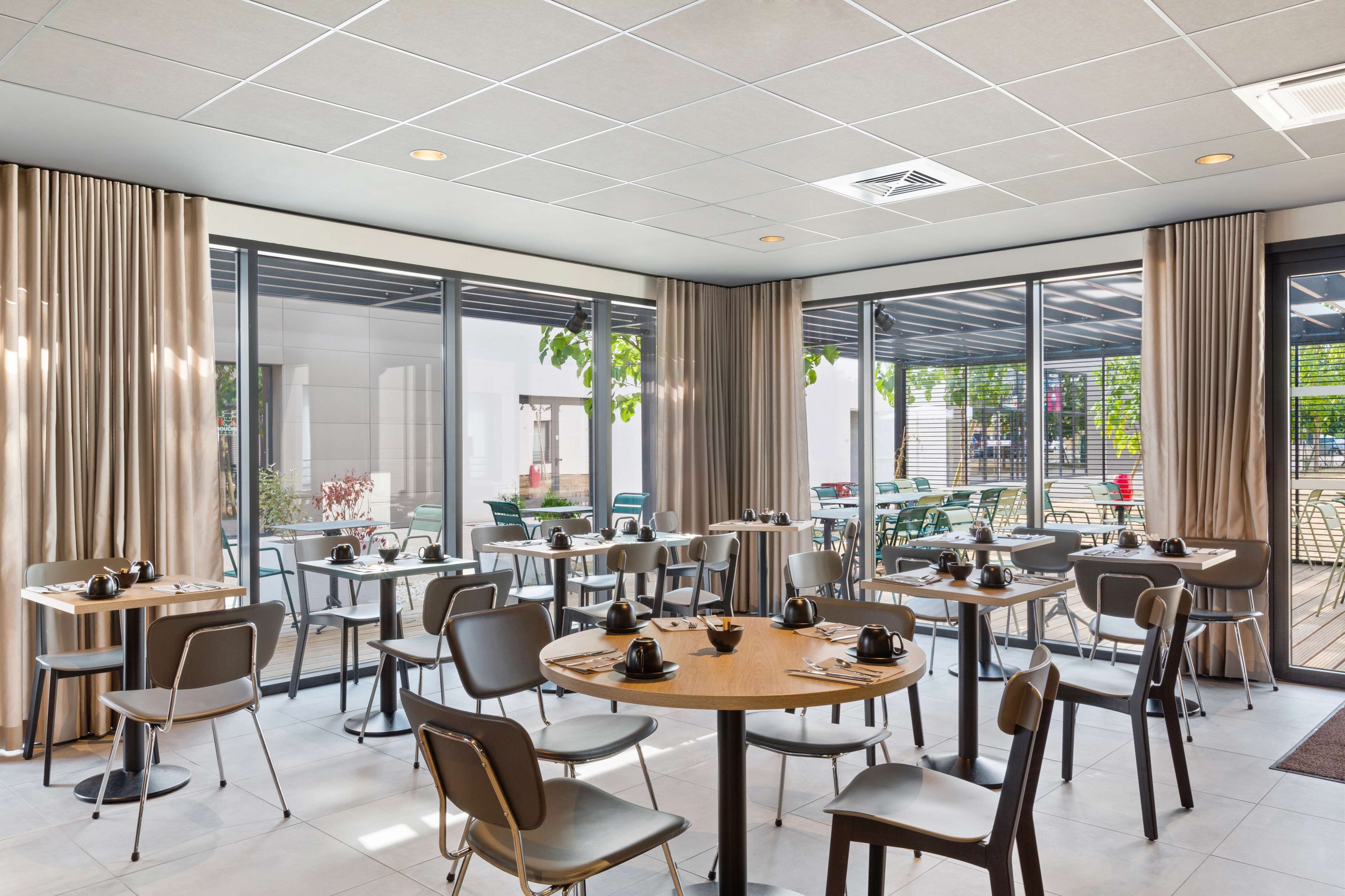 Un espace de restauration moderne avec des tables et des chaises rondes, de grandes fenêtres avec des rideaux et des sièges extérieurs visibles à travers la vitre.