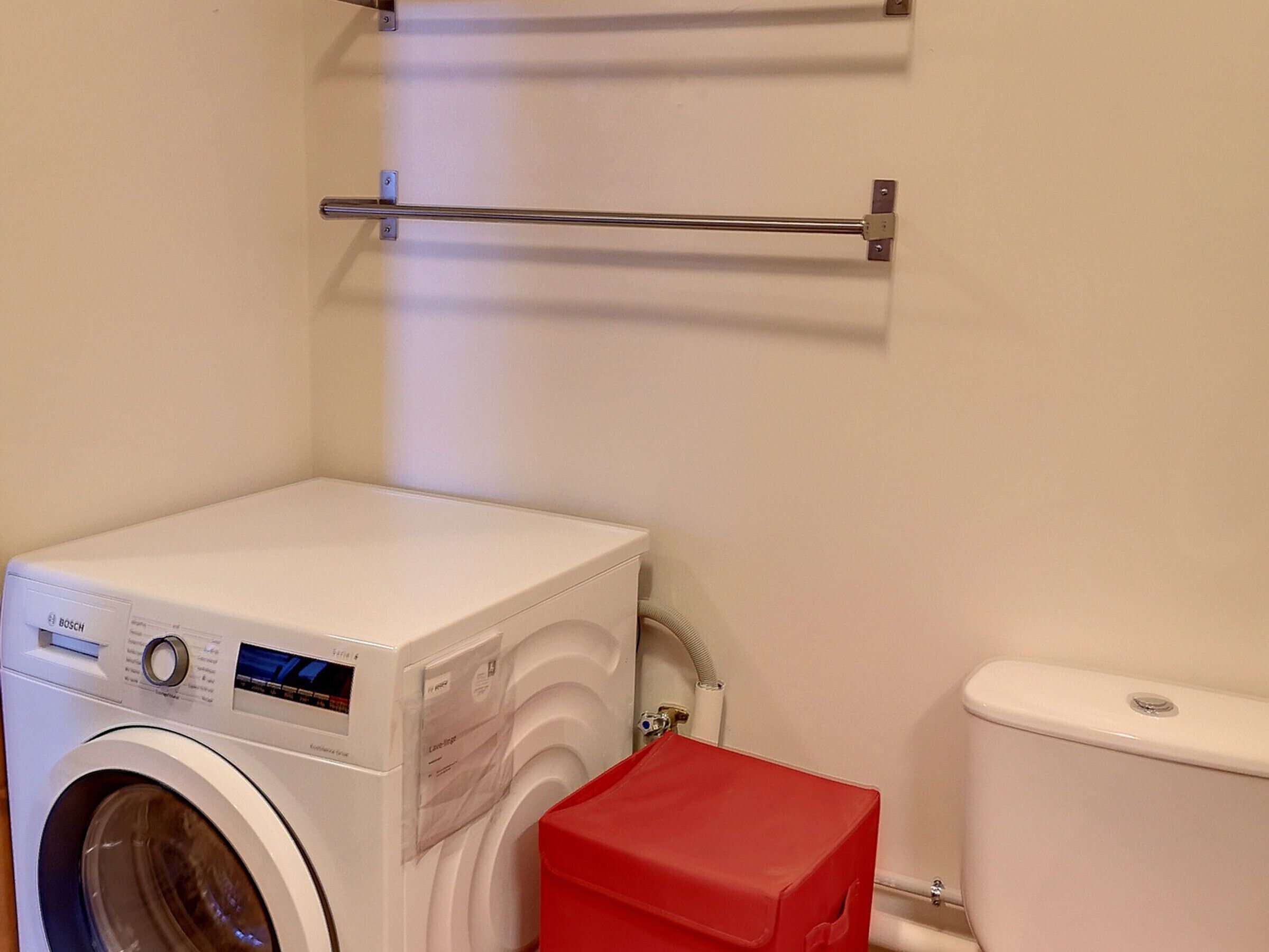 A compact, white washing machine with a control panel, a red storage box, and a metal towel rack on the wall.