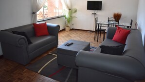 Uma sala de estar com dois sofás cinzas, um pequeno banquinho, uma televisão na parede, uma mesa de jantar com cadeiras e um vaso de planta.