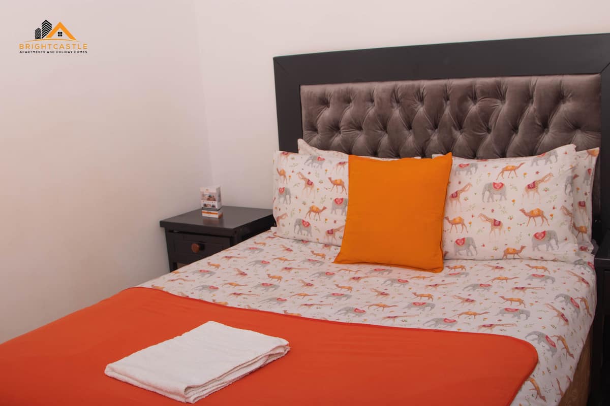 A neatly made bed with an orange bedspread and a white towel, a tufted headboard, and patterned pillows.