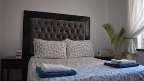 Cama bem arrumada com roupa de cama estampada, cabeceira escura e uma planta na mesinha de cabeceira.