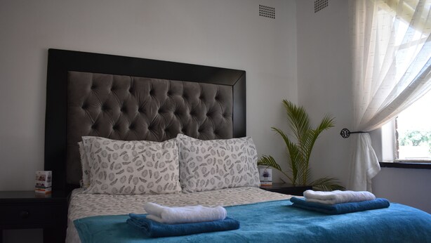 Quarto com cabeceira estofada, almofadas estampadas, colcha azul e cama bem arrumada com toalhas dobradas.