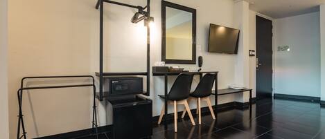 A modern hotel room with a black-framed mirror, a flat-screen TV, a desk with two black chairs, a microwave, and a small refrigerator.