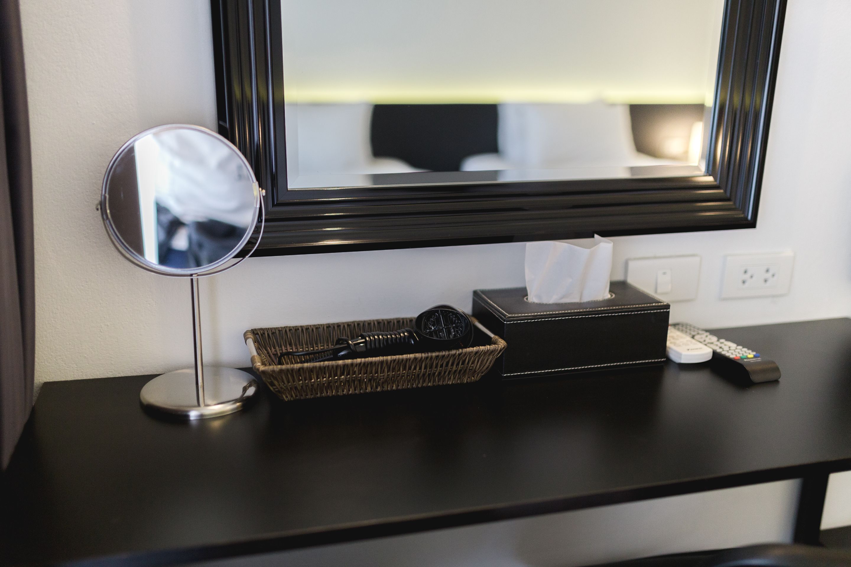 A hotel room with a black console table, a round mirror, a tissue box, a remote control, and a wicker basket.