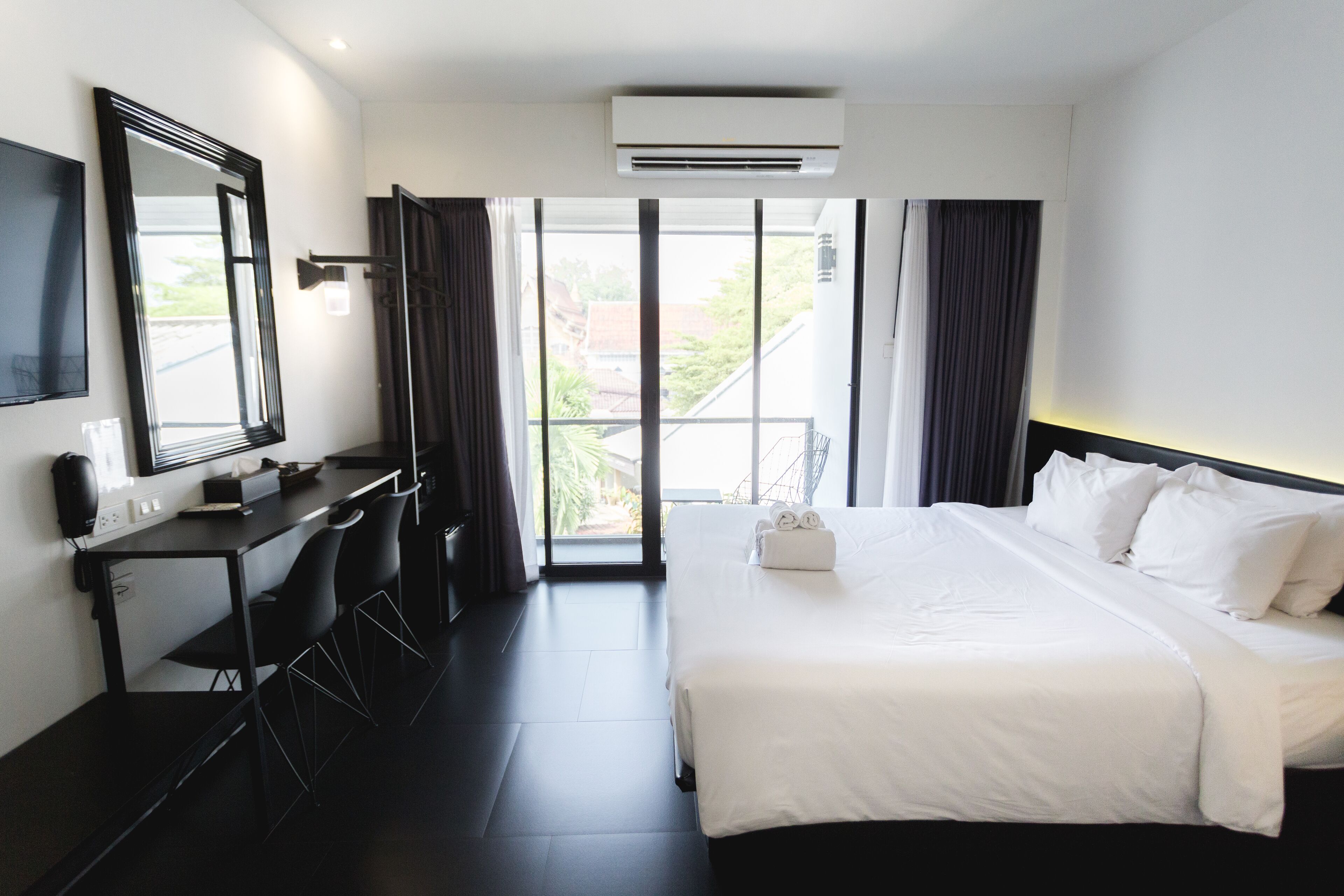 A hotel room with a bed, a desk with chairs, a TV, and a large window with curtains.