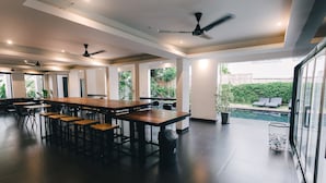 A spacious indoor area with a long wooden table and multiple chairs, ceiling fans, and large windows offering views of a garden and pool. - iSilver Hotel (Chiang Mai)