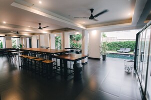 A spacious indoor area with a long wooden table and multiple chairs, ceiling fans, and large windows offering views of a garden and pool.