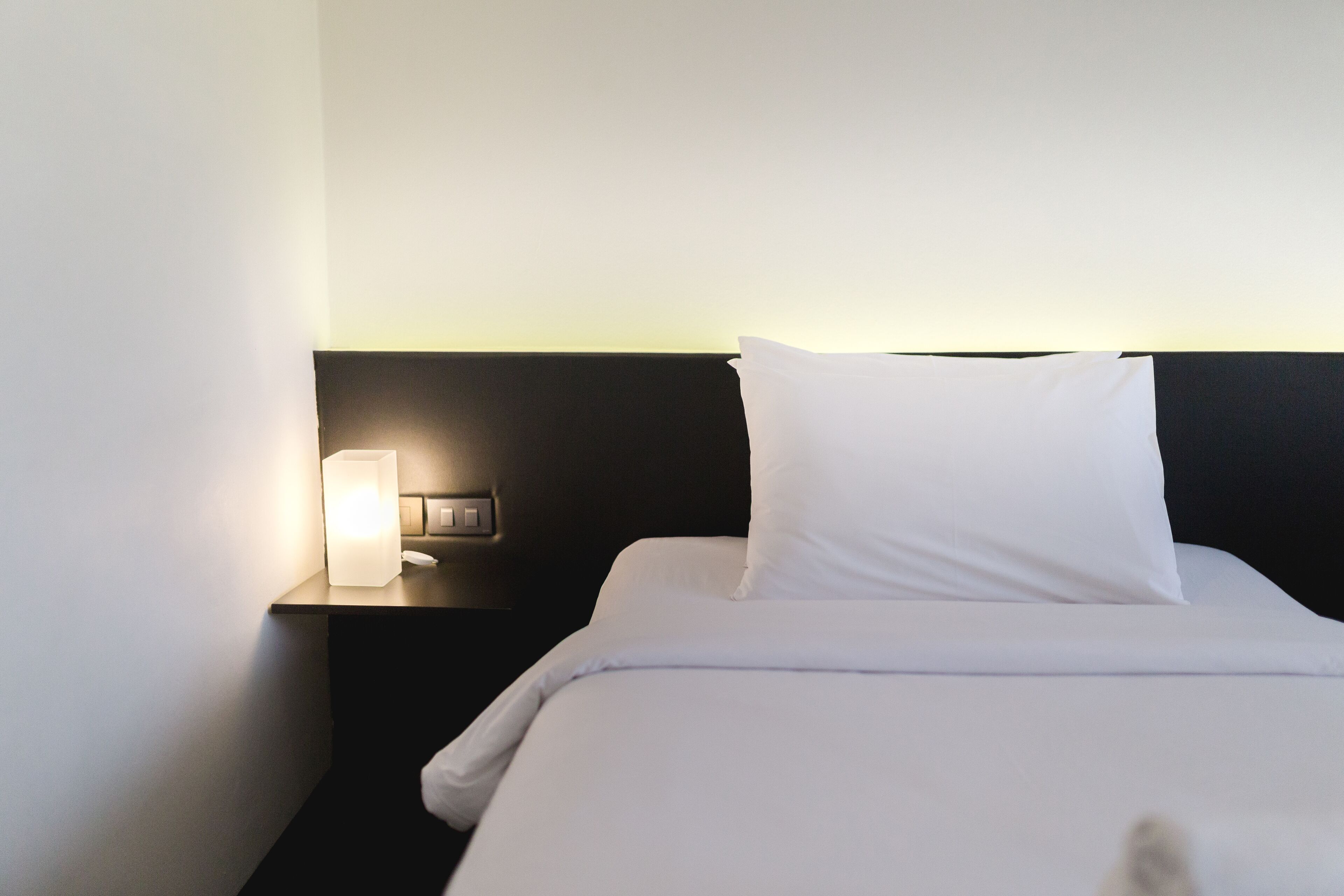 A neatly made bed with white bedding and pillows, a bedside lamp, and a dark headboard.