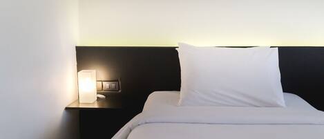 A neatly made bed with white bedding and pillows, a bedside lamp, and a dark headboard.
