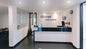 A reception area with a sign reading "iSilver Less is more," a reception desk, a clock, and a television screen. - iSilver Hotel (Chiang Mai)