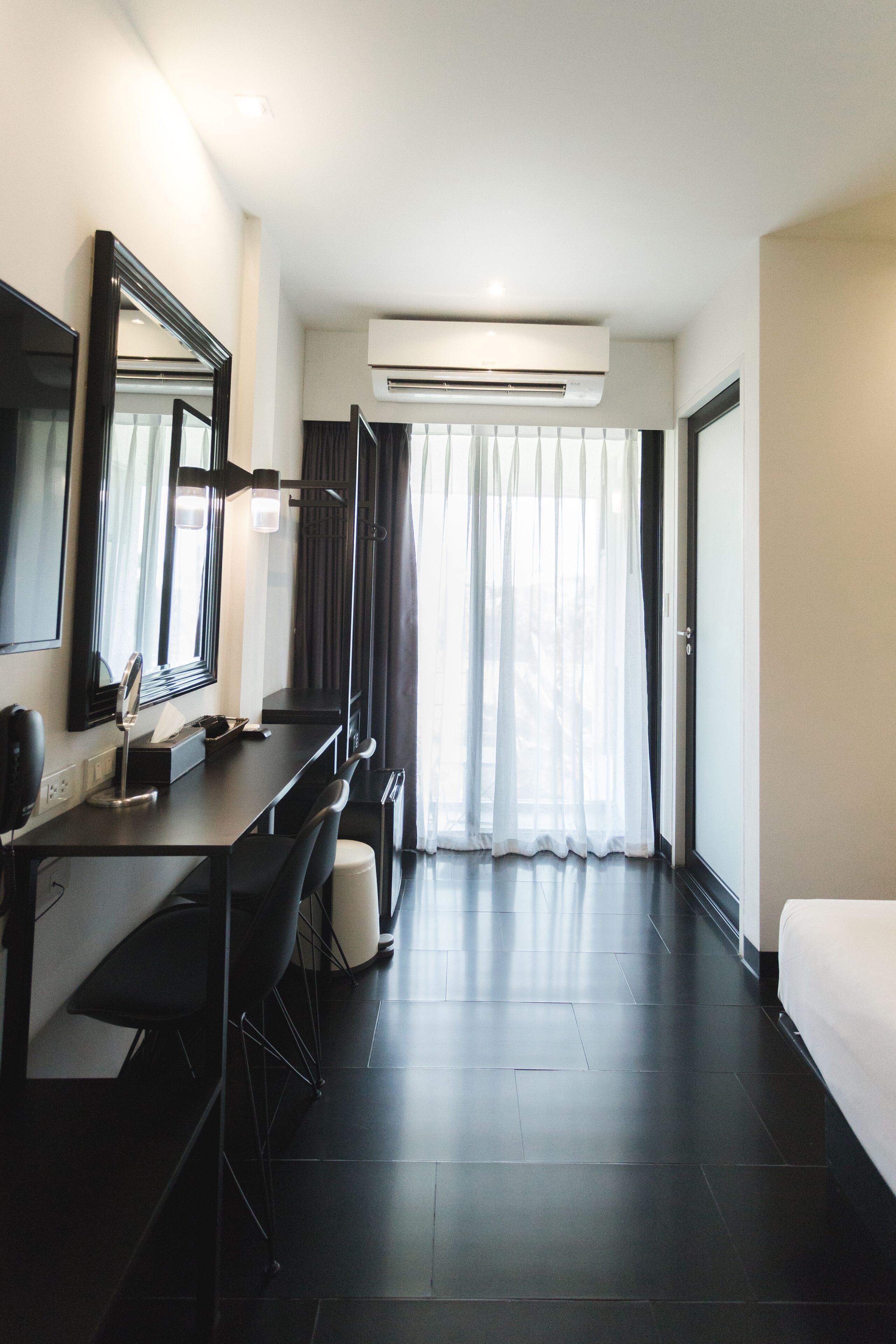 A modern hotel room with a dark wood floor, a large bed, a desk with a chair, a mirror, and a window with sheer curtains.