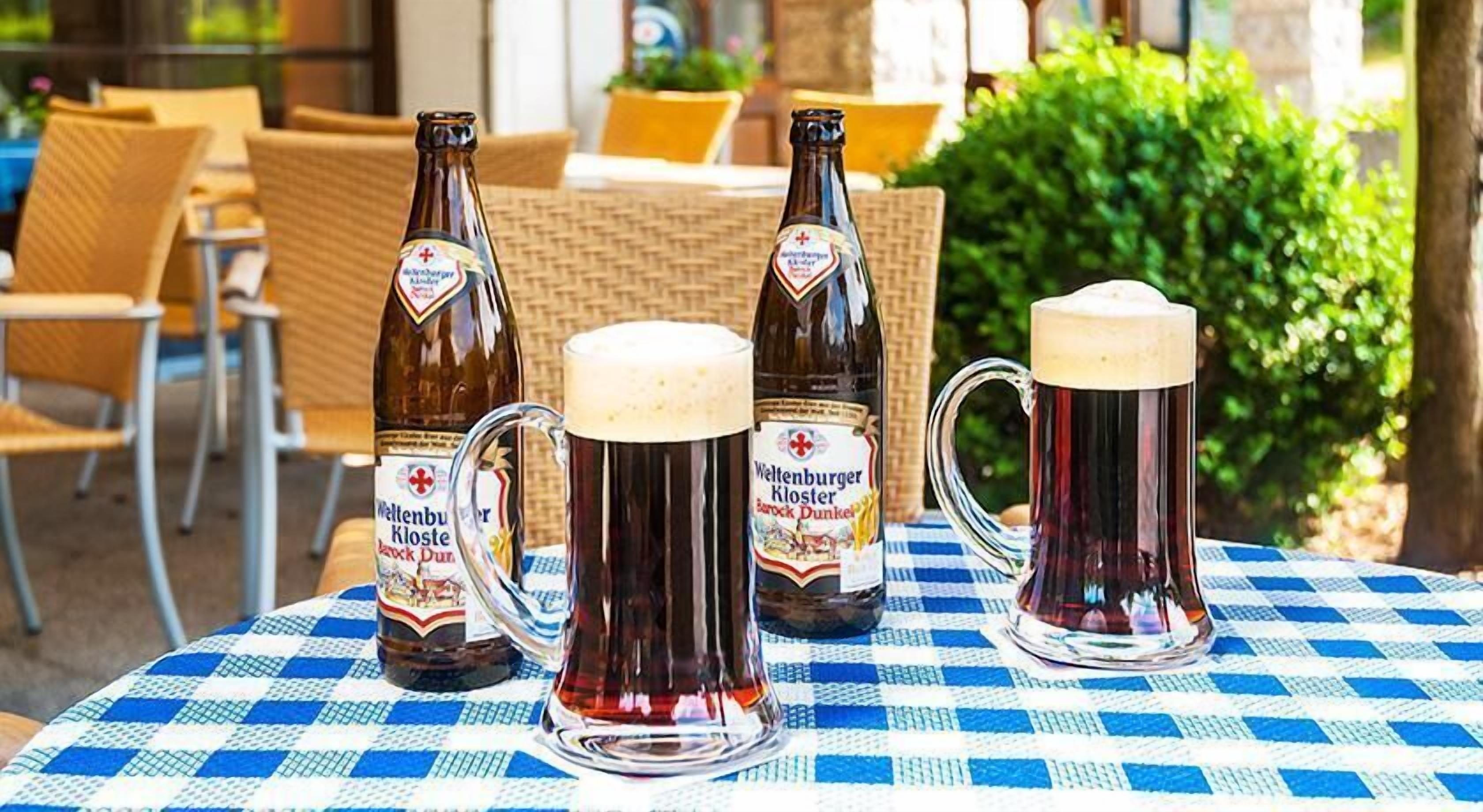 Une table avec deux bouteilles de bière et deux chopes de bière, placée à l’extérieur, avec des chaises en osier et une nappe à carreaux.