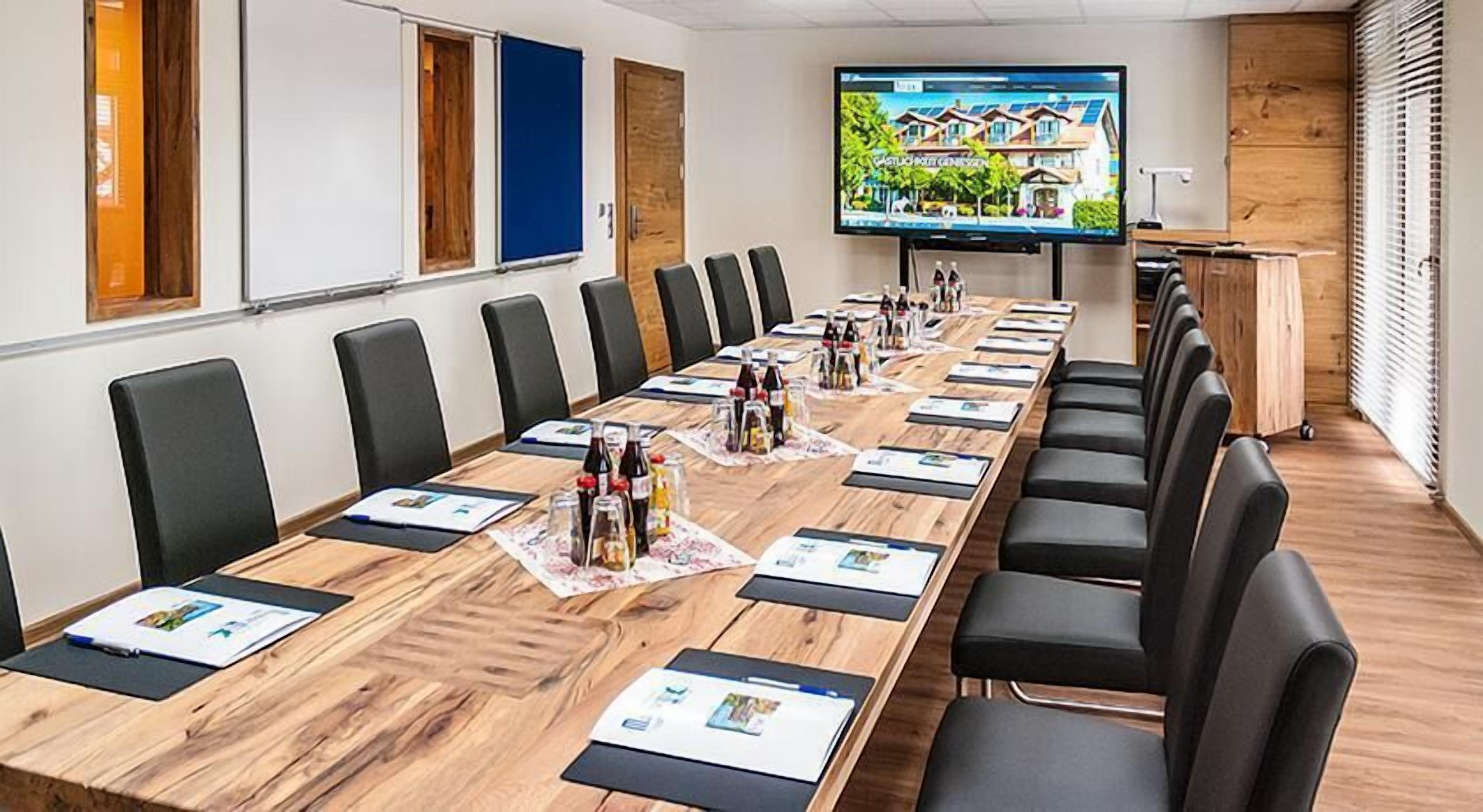 A conference room with a long table, black chairs, and a TV screen displaying a house.