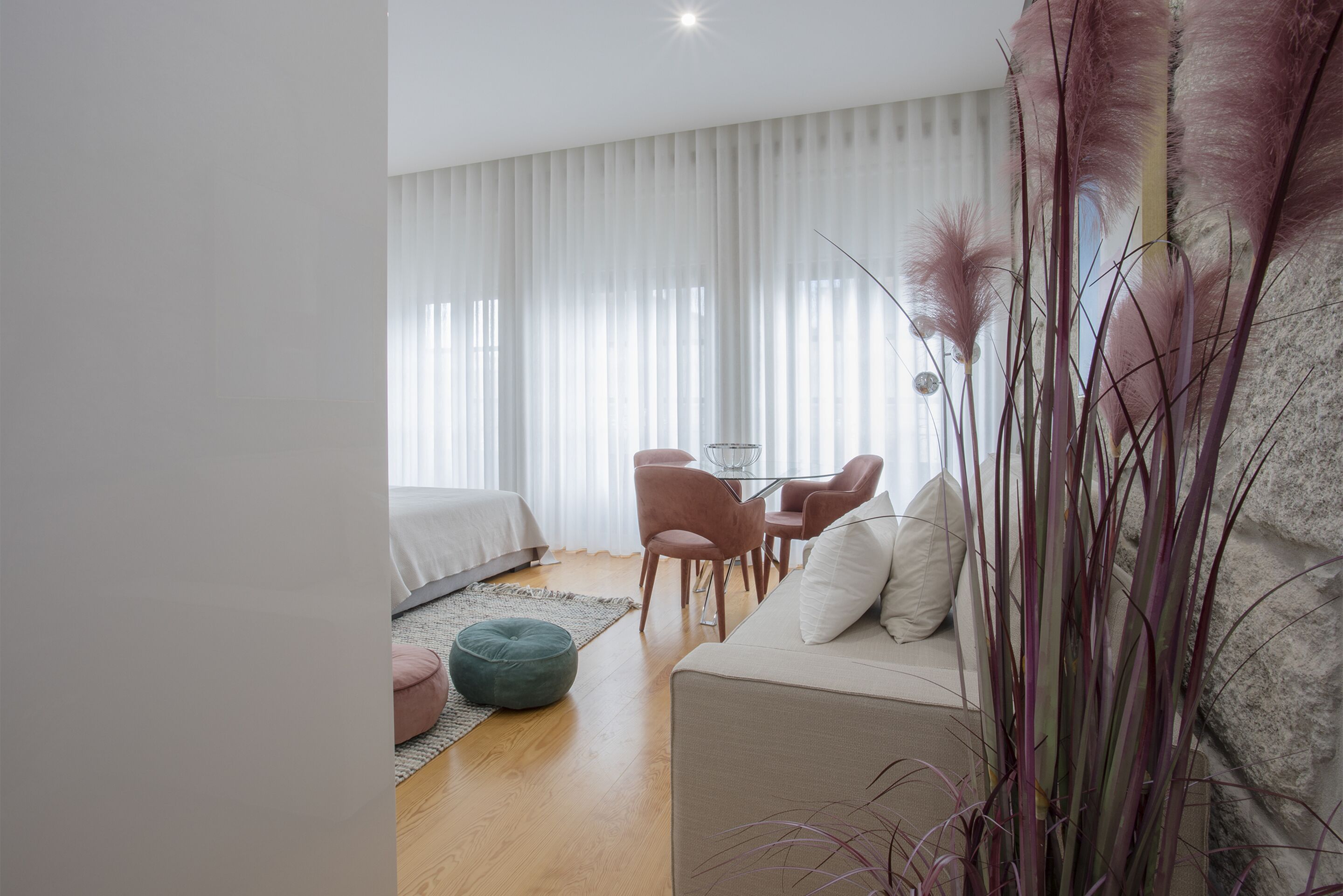 A modern bedroom with a bed, a sofa, a dining table with chairs, and a large window with sheer curtains.
