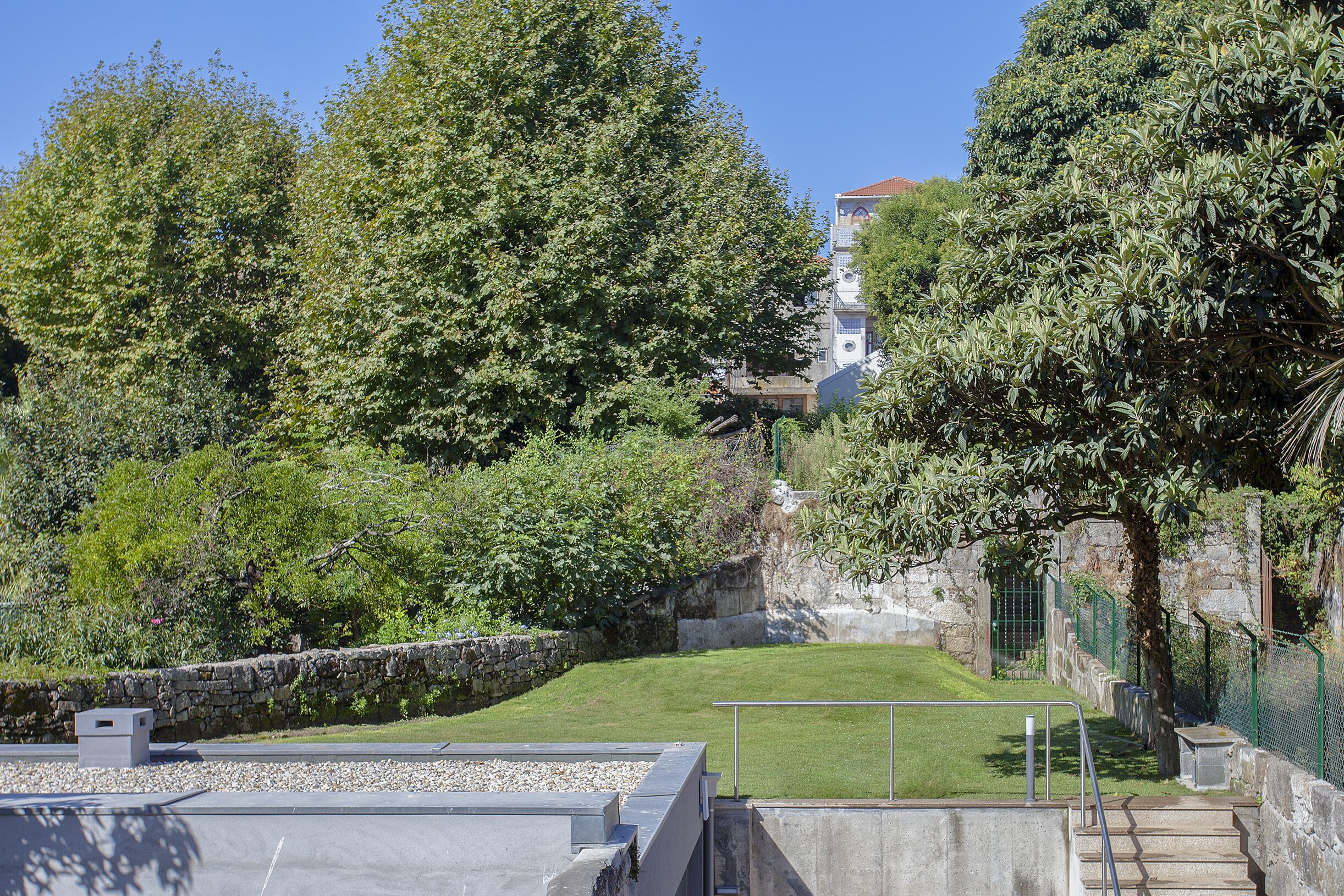 Un patio con muro de piedra, un área con césped y una escalera que lleva a un edificio con tejado rojo.