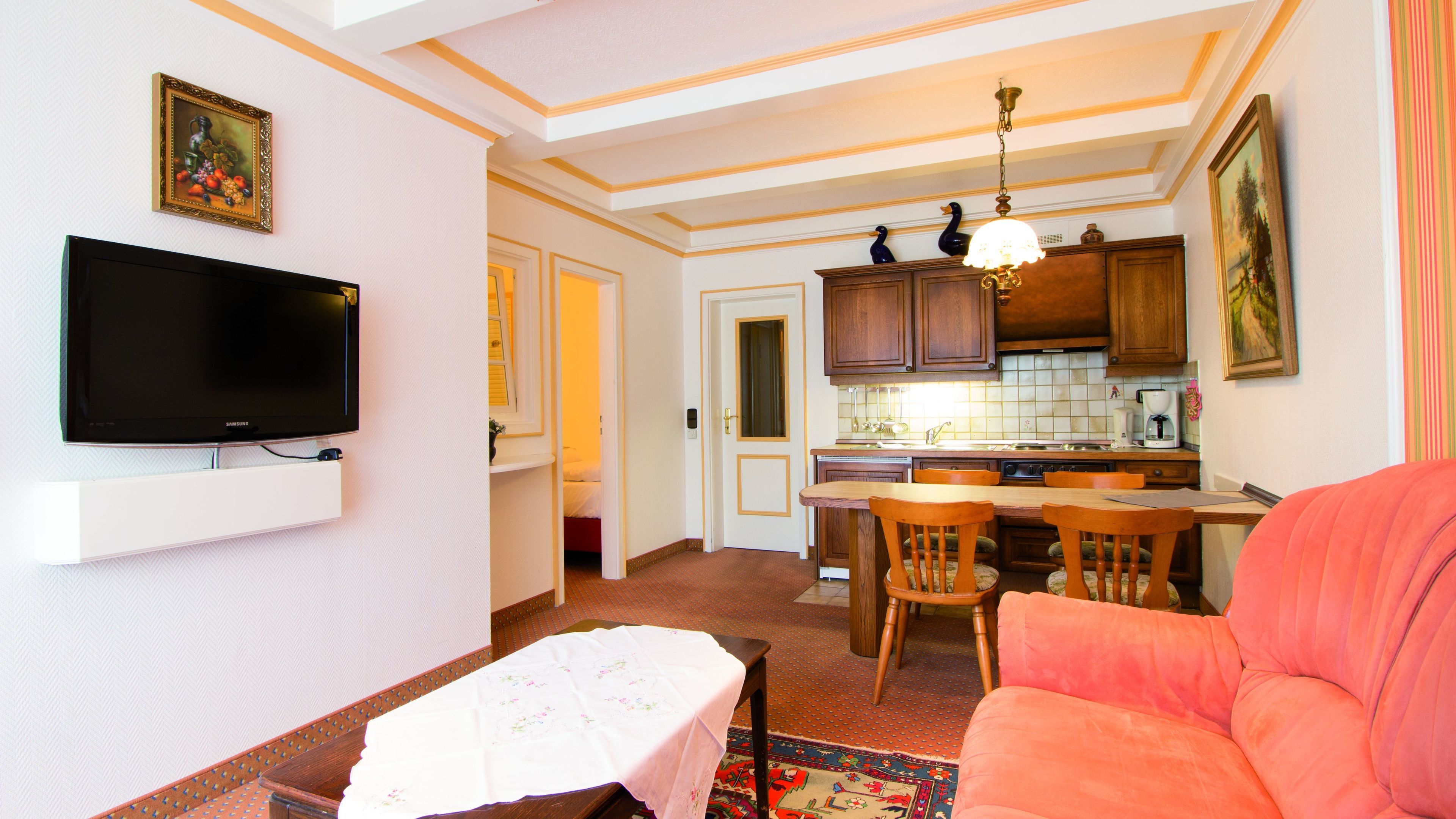 Un espacio habitable compacto con un televisor montado en la pared, una zona de comedor con sillas, una cocina con armarios de madera y una alfombra estampada en el suelo.