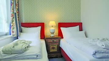 A hotel room with two single beds, a nightstand with a lamp, floral curtains, and a patterned carpet.