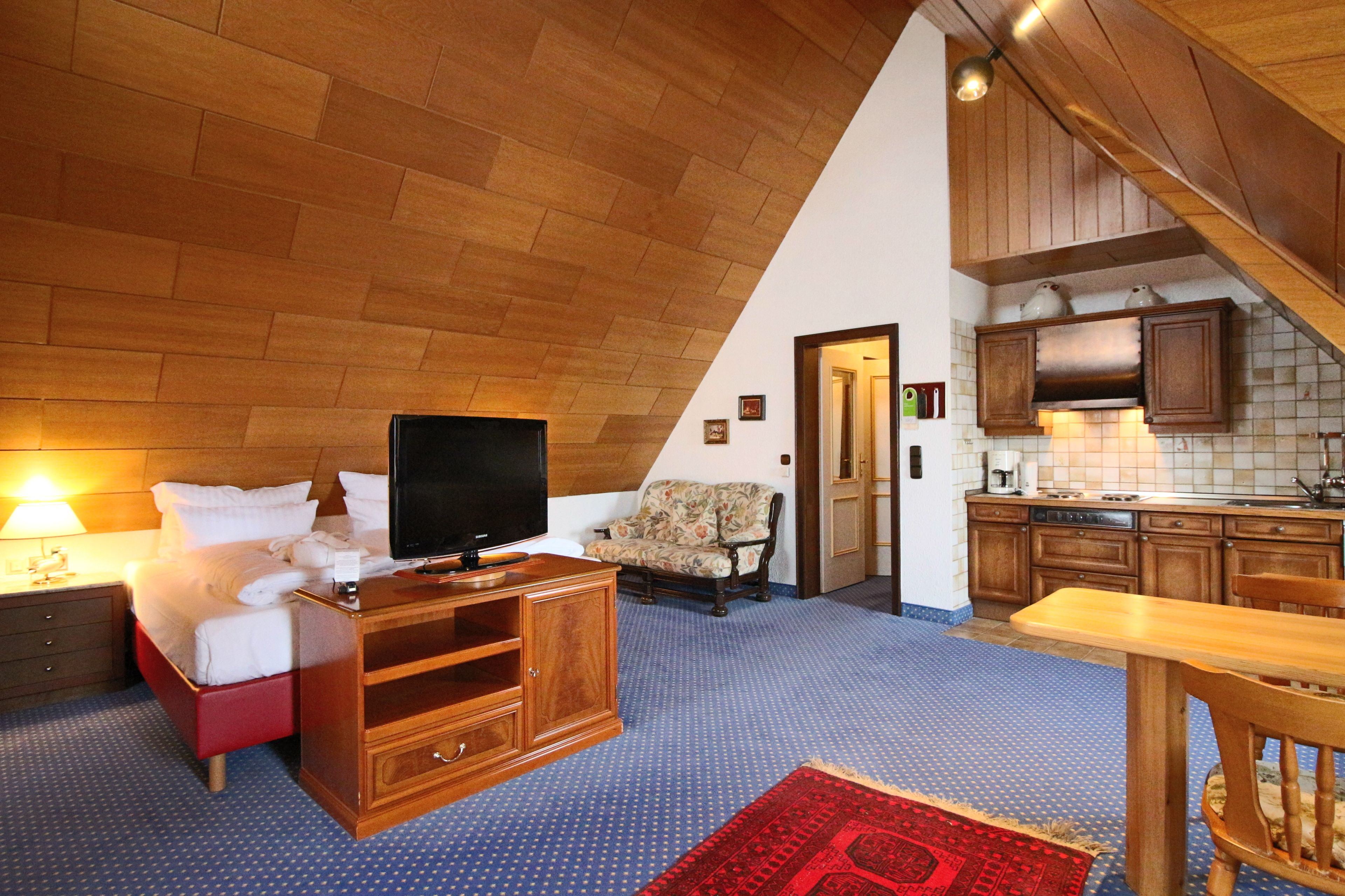 A hotel room with a wooden ceiling, a bed with white linens, a television on a wooden stand, a chair, a dining table with chairs, and a kitchen area with wooden cabinets and a sink.