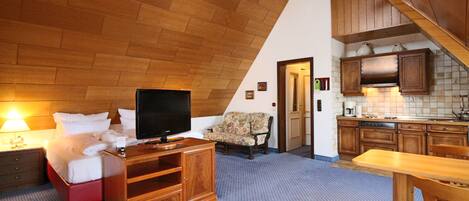 Habitación de hotel con techo de madera, cama con sábanas blancas, televisor sobre un mueble de madera, una silla, mesa de comedor con sillas y zona de cocina con armarios de madera y un lavamanos.