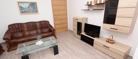 Ein modernes Wohnzimmer mit einem braunen Ledersofa, einem Glastisch mit Pflanze, einem Fernseher, Holzschränken und einem Gemälde an der Wand.