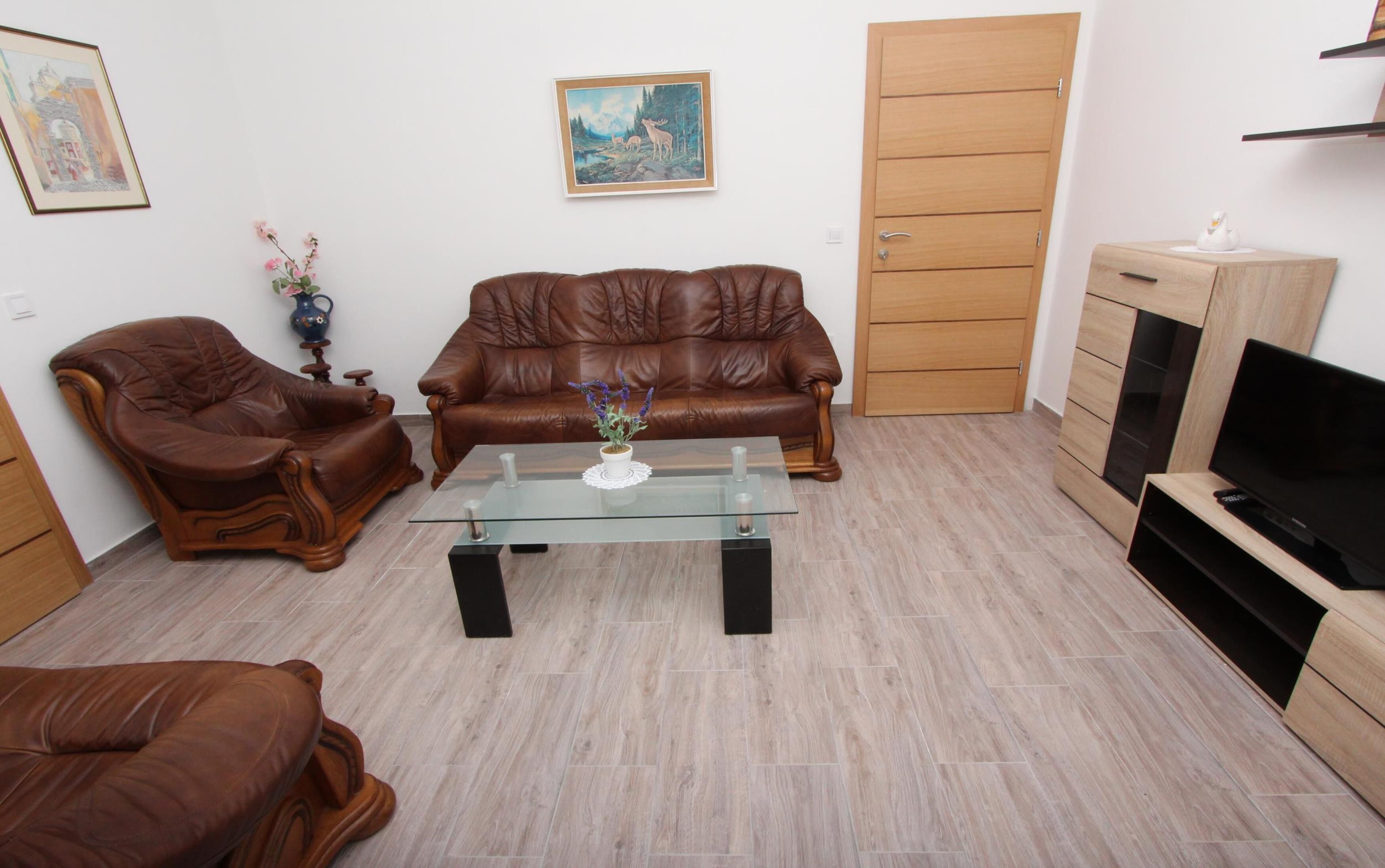 Un salon avec un canapé en cuir marron, une table basse en verre ornée d’un vase, un meuble en bois sur lequel se trouve un téléviseur, et une porte.