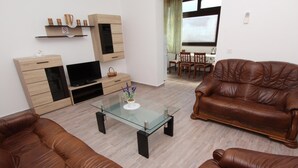 Ein Wohnzimmer mit einem Glastisch, zwei braunen Ledersofas, einem TV-Schrank und einem Essbereich im Hintergrund.