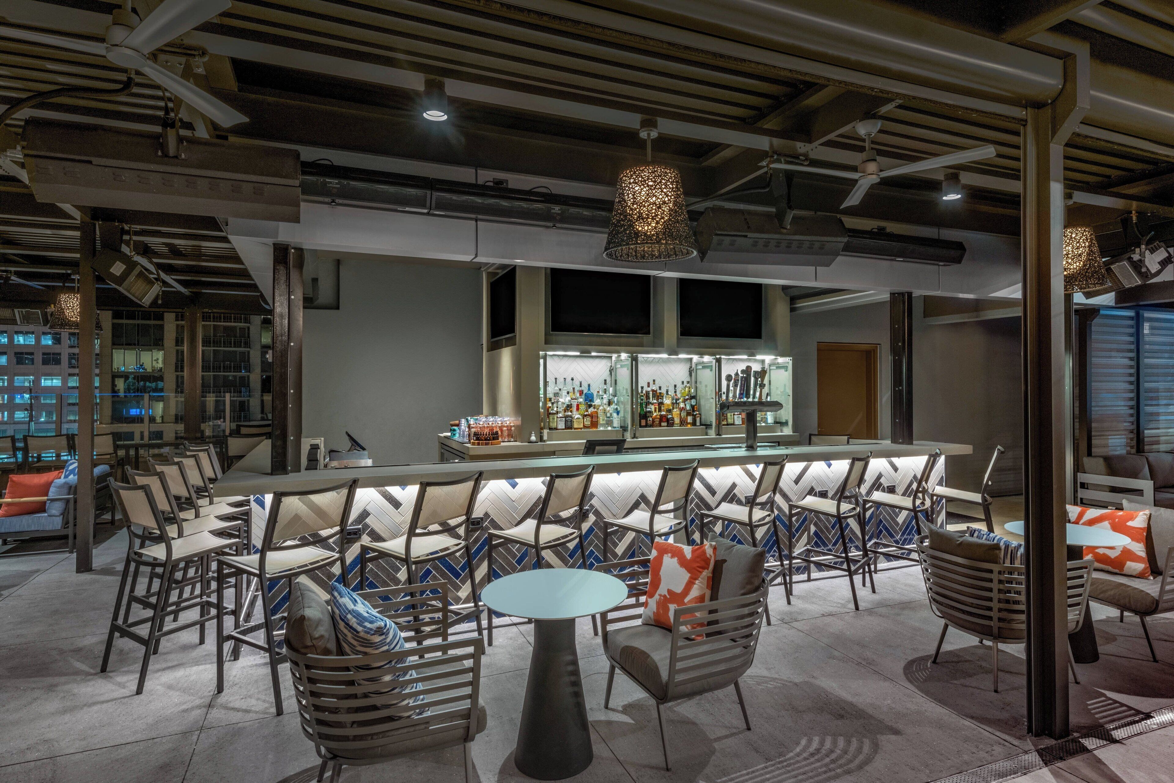 Zona de bar con una barra larga, diversas opciones de asientos que incluyen mesas redondas y sillas, y un diseño de techo moderno con vigas y luminarias a la vista.