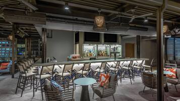 Zona de bar con una barra larga, diversas opciones de asientos que incluyen mesas redondas y sillas, y un diseño de techo moderno con vigas y luminarias a la vista.