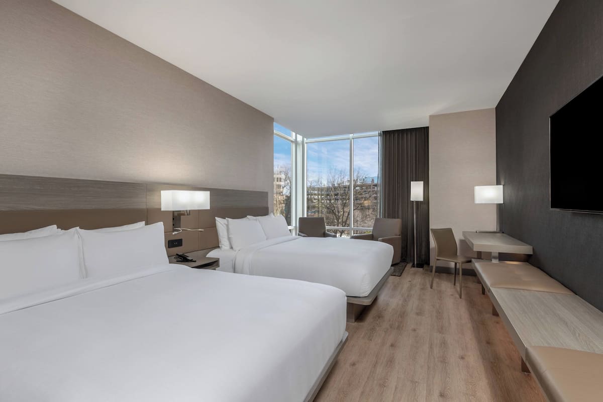 A hotel room with two beds, a desk, a chair, a TV, and a window with a city view.