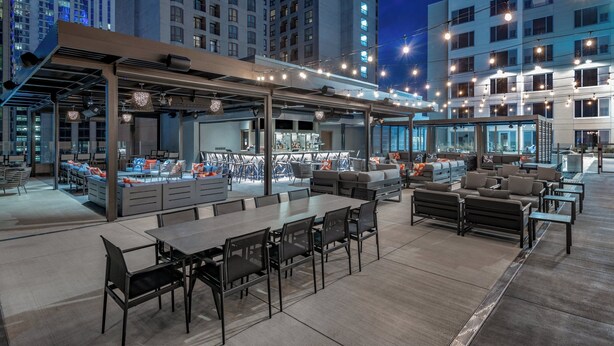 Un área de comedor al aire libre con mesas y sillas, un espacio de bar con asientos y vistas a edificios altos en el fondo.