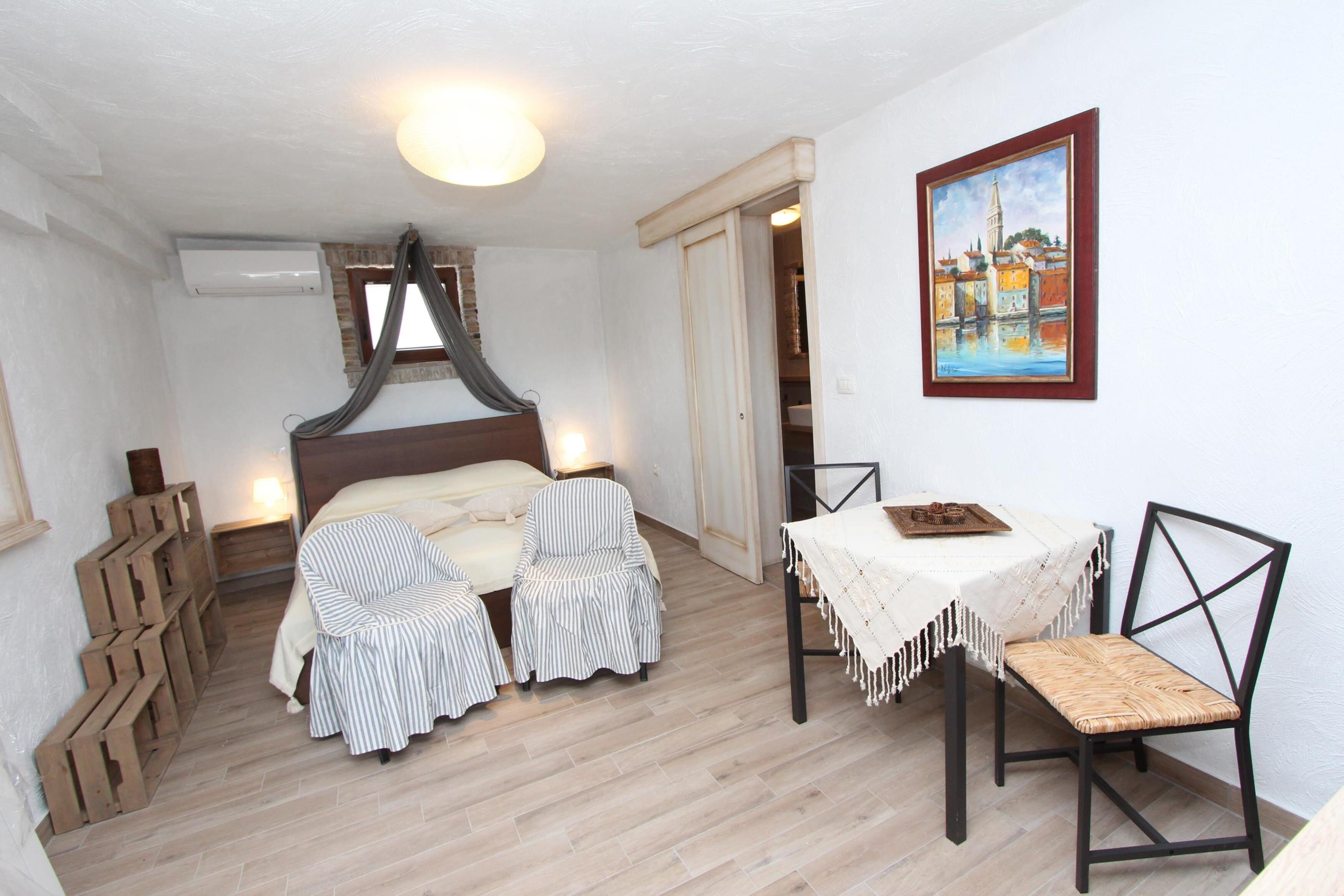 A bedroom with a bed, two striped chairs, a table with a white tablecloth, and a painting on the wall.