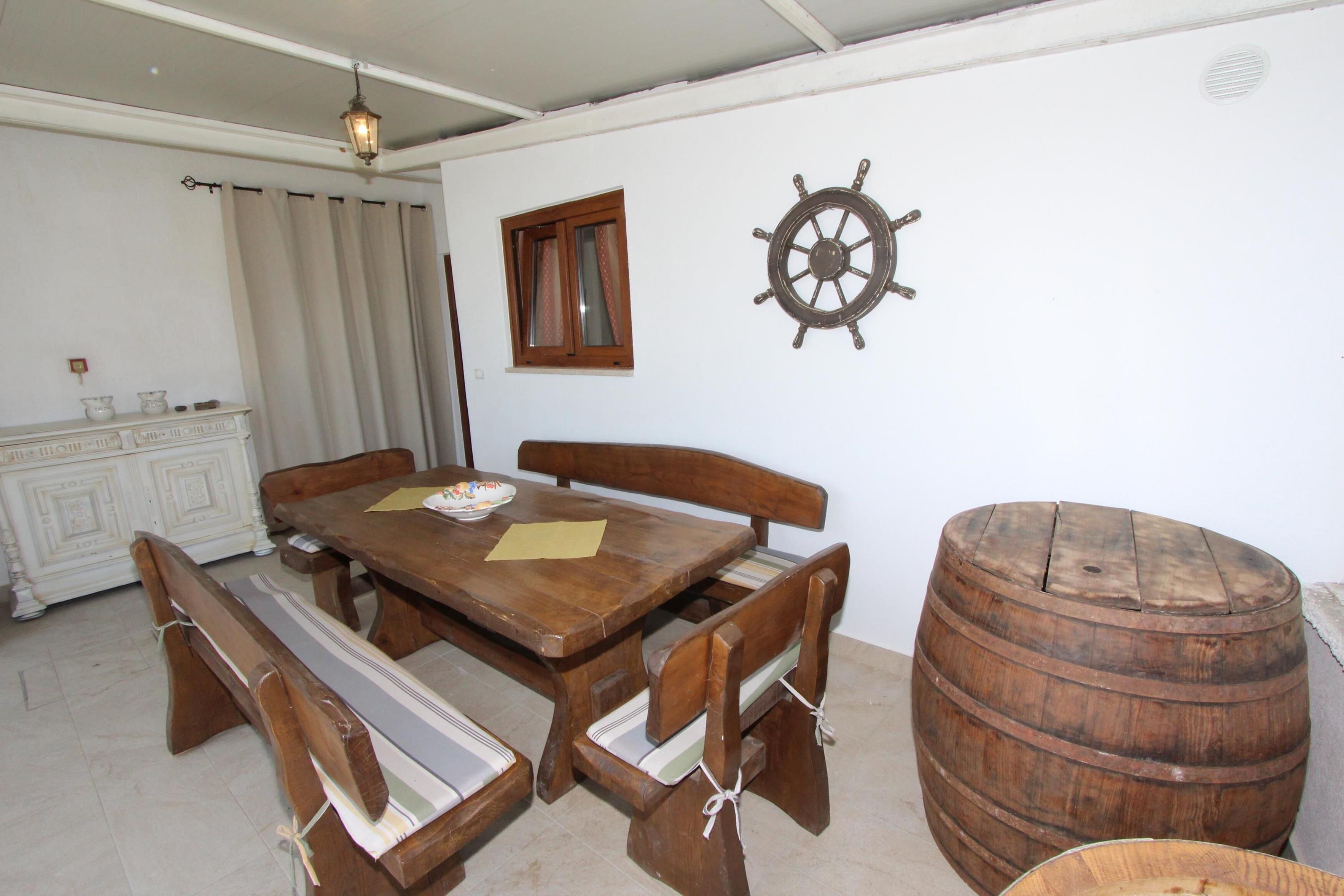 A wooden dining table with benches, a ship's wheel decoration, a wooden barrel, and a window with curtains.
