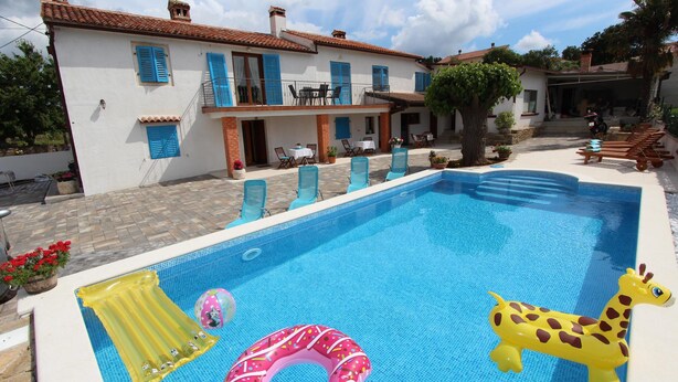 Une piscine avec des jeux gonflables, un espace extérieur avec des chaises et des tables, et un bâtiment aux volets bleus.