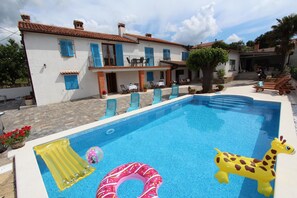 Una piscina con juguetes inflables, un área de patio con sillas y mesas, y un edificio con contraventanas azules.