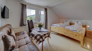 A bedroom with a wooden bed, a sofa, a table with a checkered tablecloth, a chair, and a window with curtains. - Gasthaus Zur Einkehr (Neureichenau)