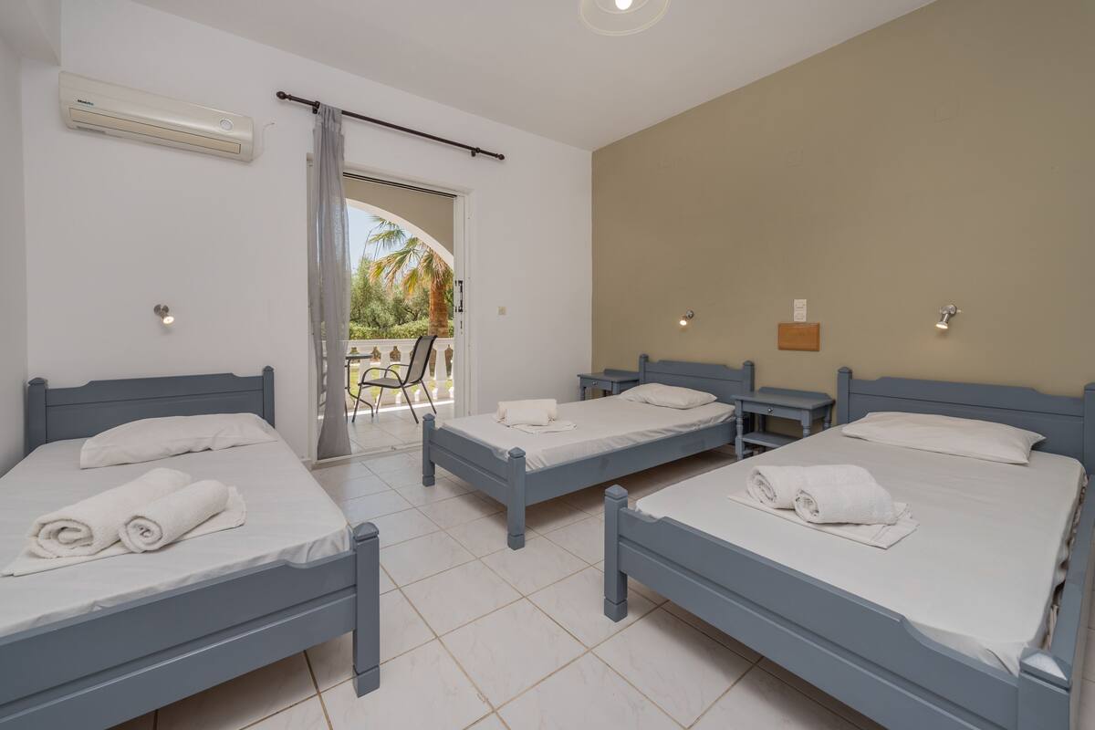 A hotel room with three single beds, a balcony with a view of palm trees, and an air conditioning unit on the wall.