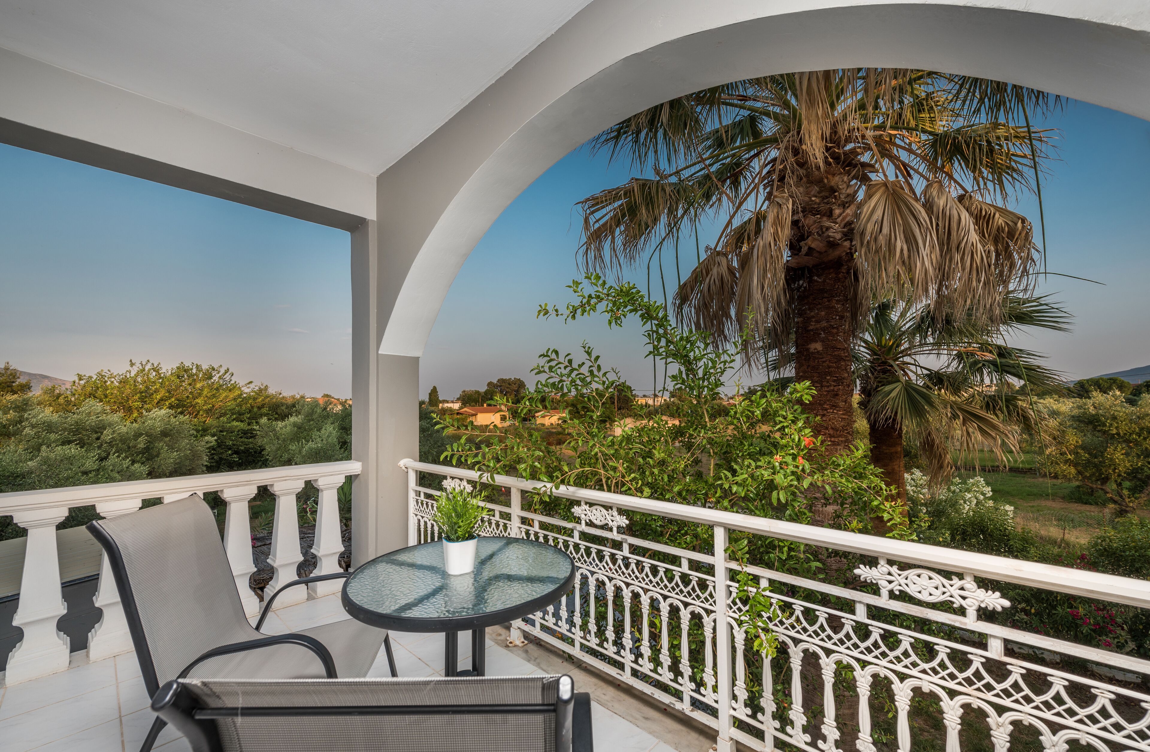 A balcony with a round table and chairs, white balustrade, and a view of a palm tree and greenery.