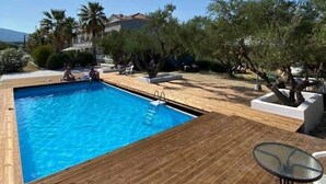 Une piscine dotée d’une terrasse en bois, entourée d’arbres et avec un bâtiment visible au loin.