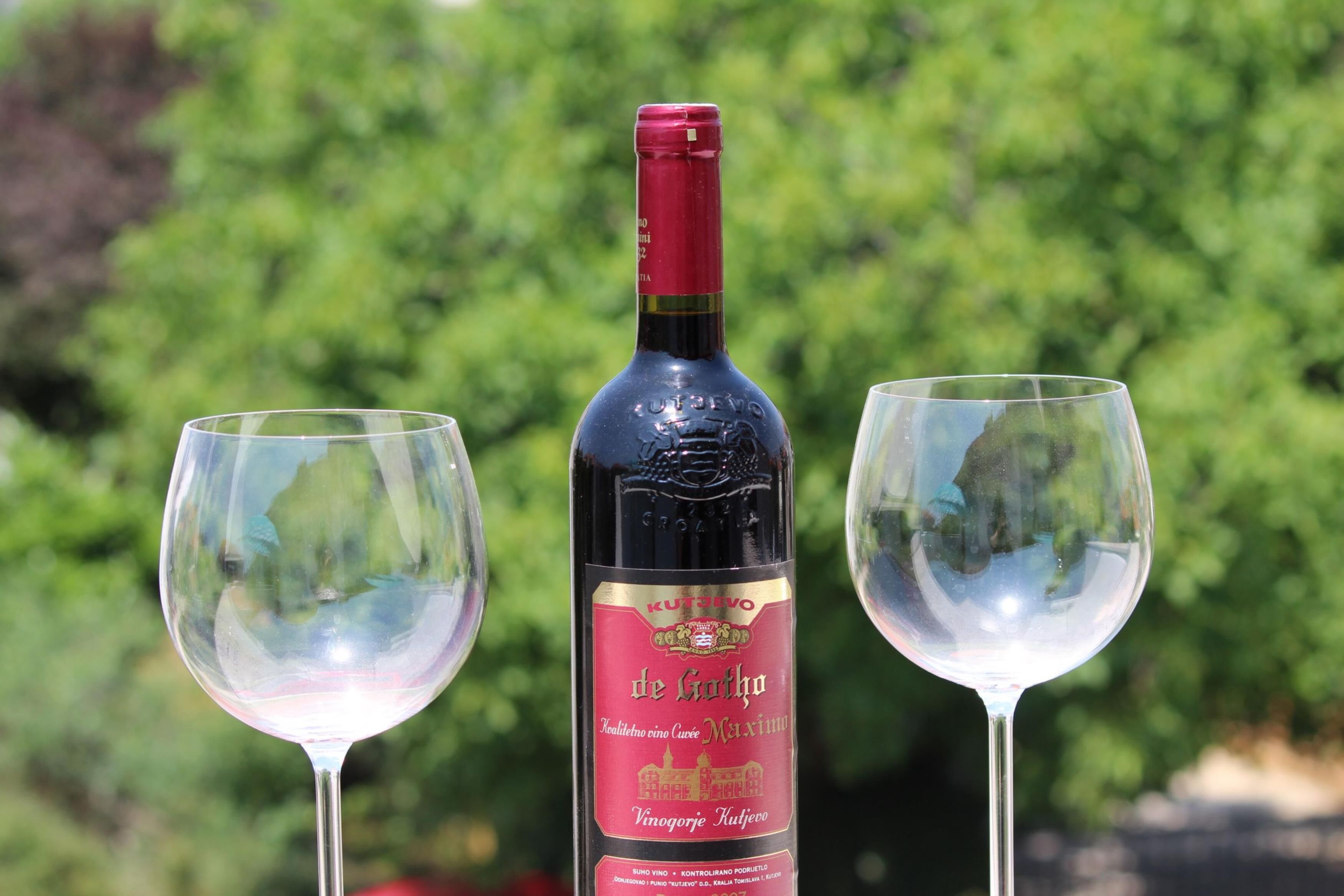 A bottle of wine with a label, two empty wine glasses, and a blurred green background.