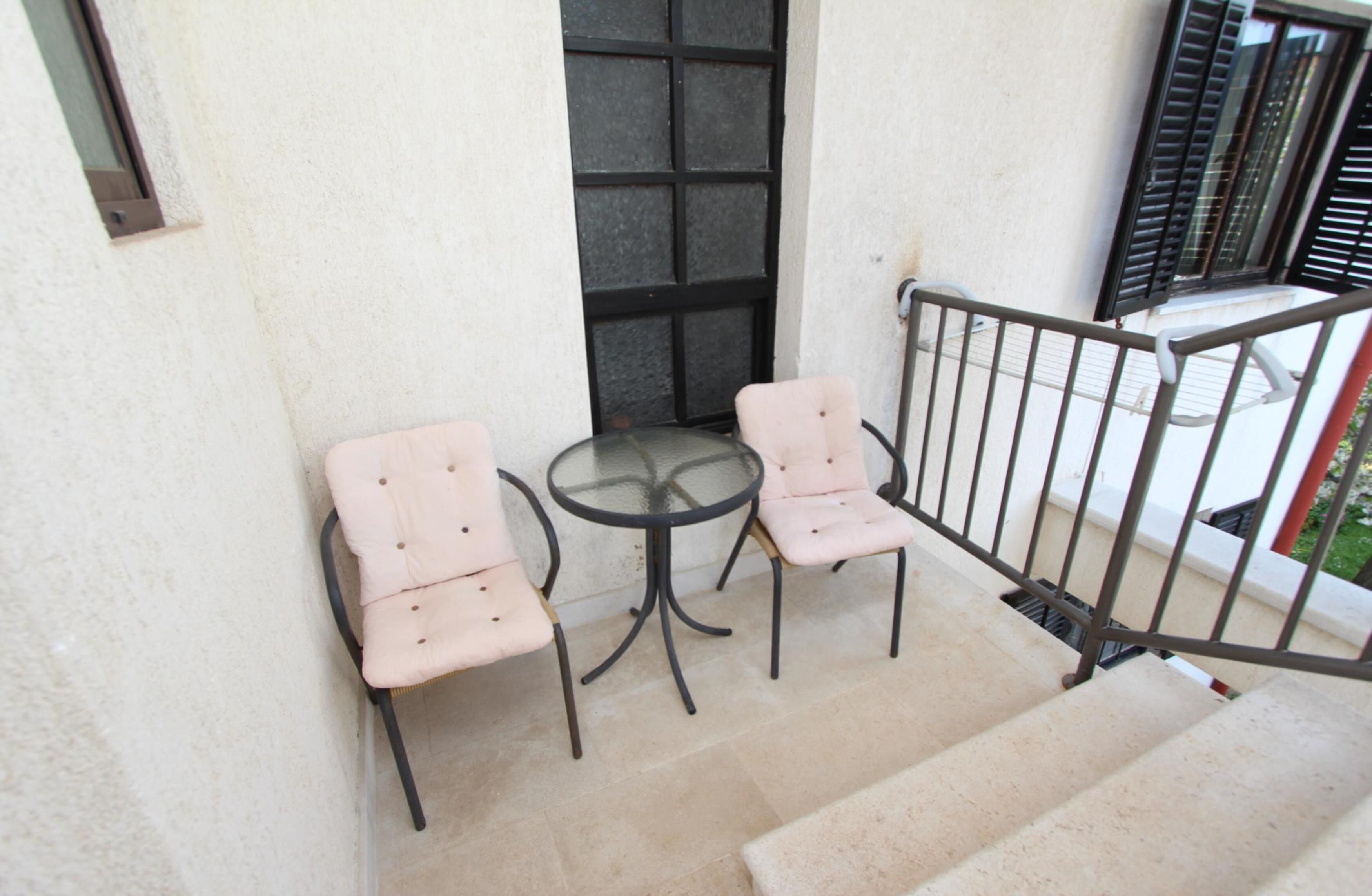 Ein kleiner Außenbereich mit zwei rosa gepolsterten Stühlen und einem Glastisch, auf einem Balkon mit Metallgeländer und Blick auf den Garten.