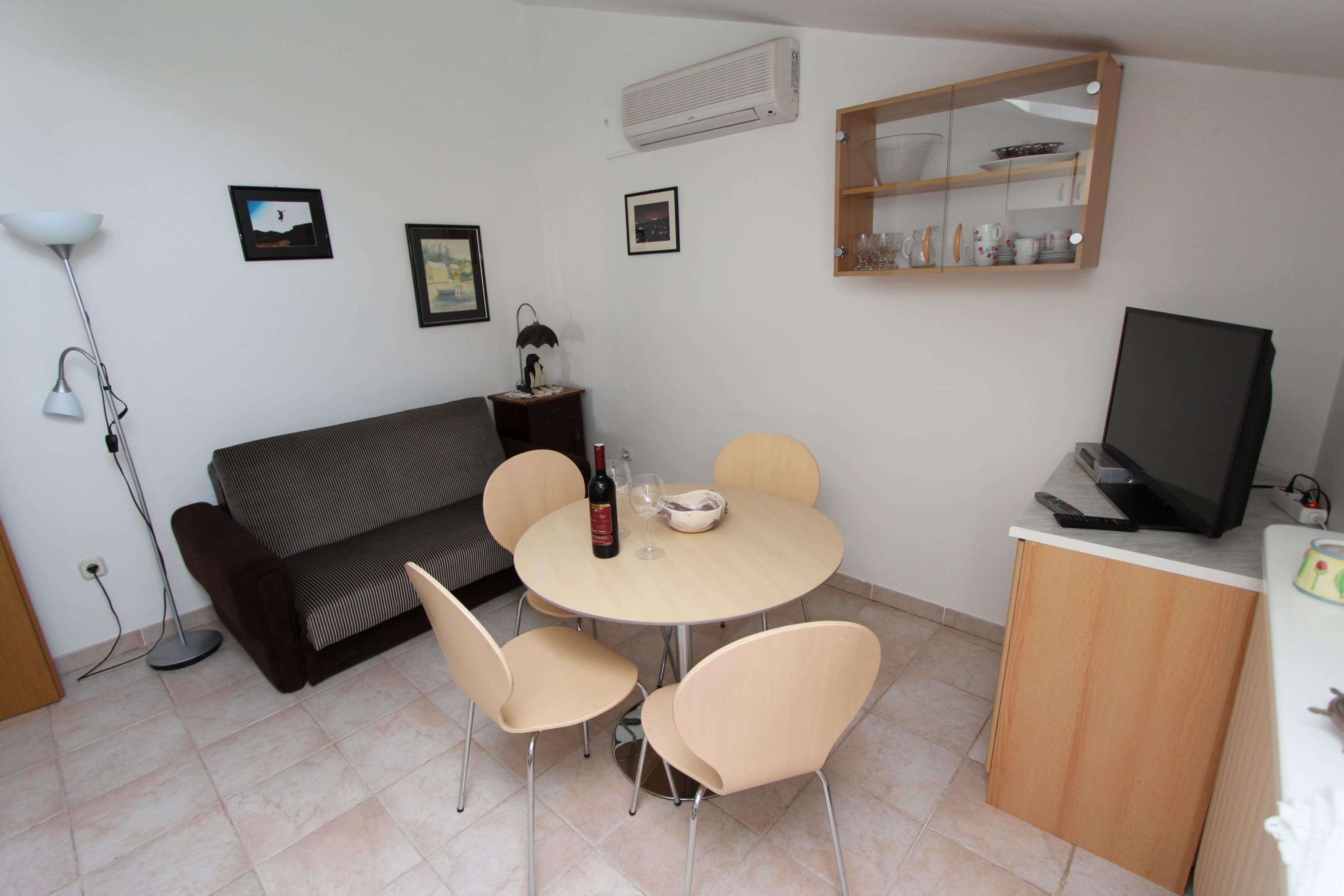 A compact living space with a sofa, a round dining table with four chairs, a television on a stand, and a cabinet with glass doors.