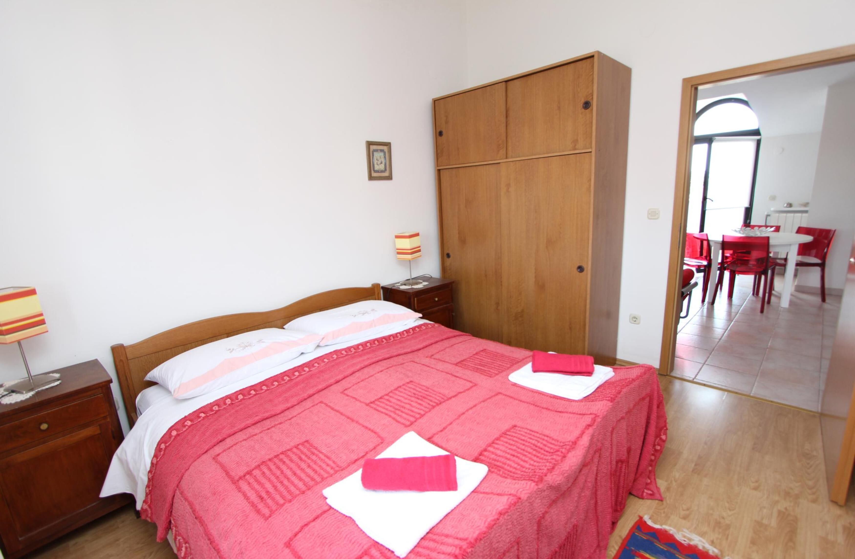 A bedroom with a wooden wardrobe, a bed with a red and white bedspread, a nightstand with a lamp, and a mirror reflecting a dining area with red chairs.