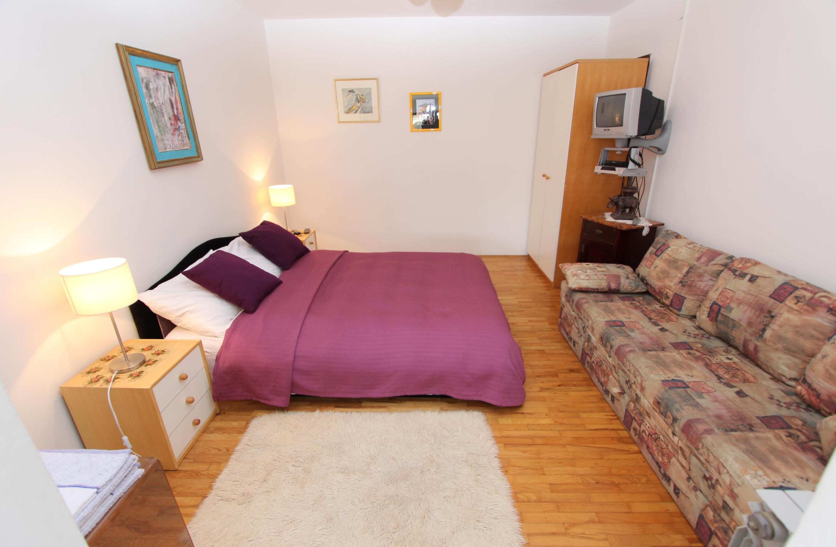 A small, cozy bedroom with a bed, a sofa, a TV, and a wooden nightstand.