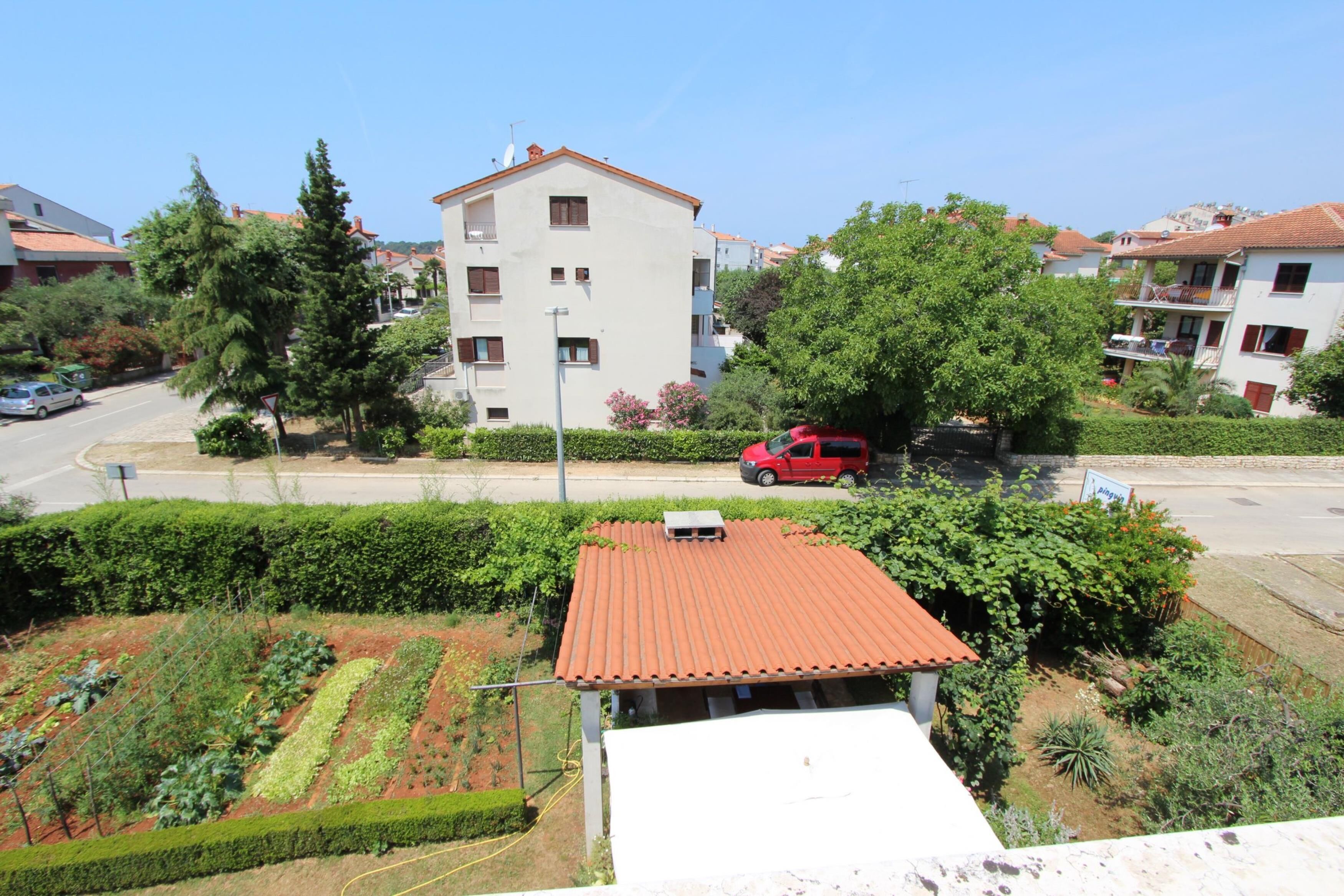 Un quartiere residenziale con un edificio bianco dotato di tetto rosso, immerso nel verde e affiancato da una strada con un'auto rossa parcheggiata.