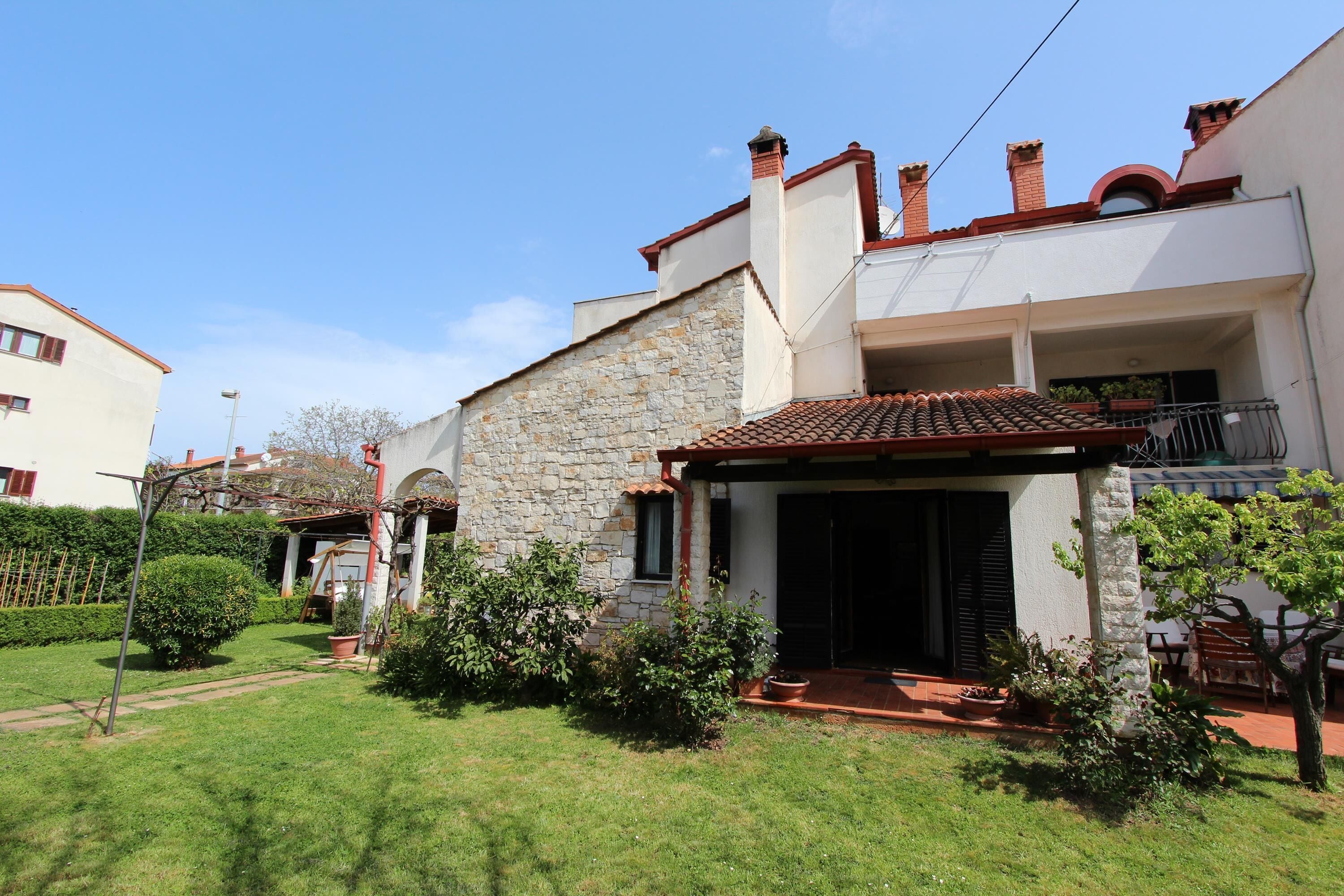 A two-story house with a stone facade, a tiled roof, and a small garden with a pathway leading to the entrance.