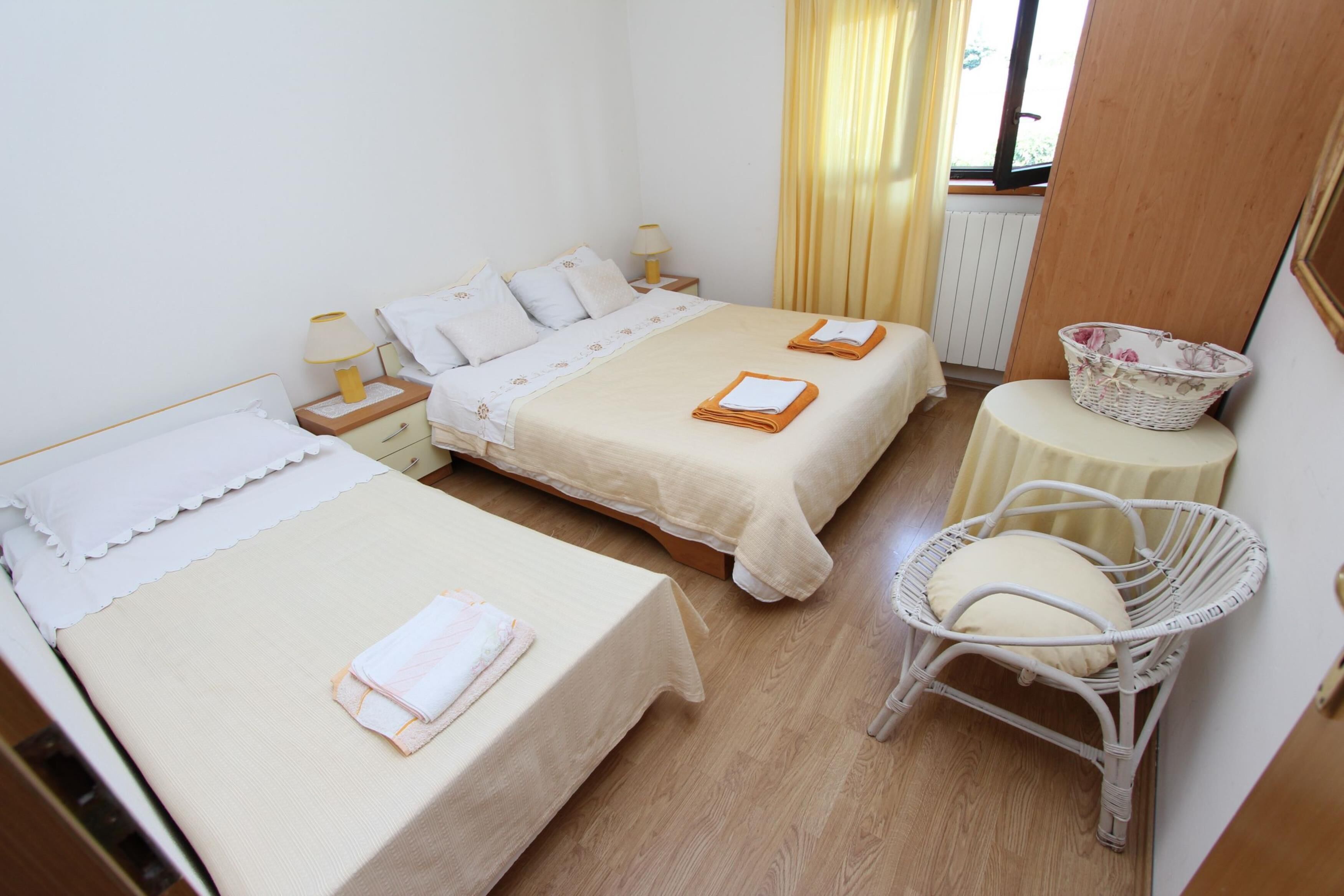 Quarto de hotel com duas camas, uma cadeira de vime, uma mesa pequena e uma janela com cortinas.
