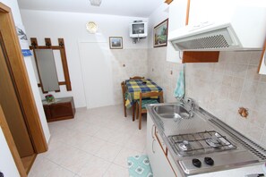 A compact kitchen with a gas stove, sink, and tiled backsplash. There is a dining table with a checkered tablecloth and chairs. A small cabinet and a mirror are visible. - Apartments Milka (Rovinj)